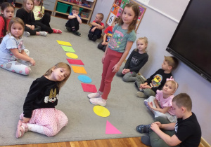 Zdjęcie przedstawia dzieci i dziecko spacerujące po dywanie utworzonym z figur geometrycznych.