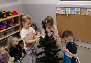 Troje dzieci stojących wokoło choinki wieszają na niej bombki.