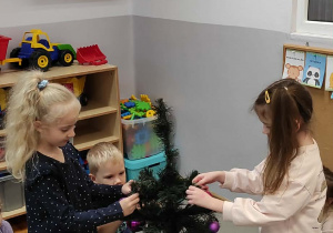 Dwie dziewczynki oraz chłopiec przyozdabiają bombkami grupową choinkę.