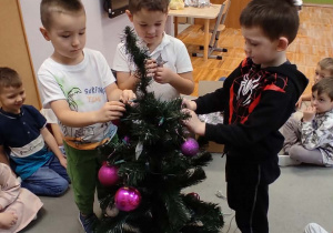 Trzej chłopcy stojący wokół choinki wieszają na niej bombki.