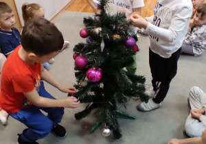 Trzej chłopcy stojący wokół choinki wieszają na niej bombki.