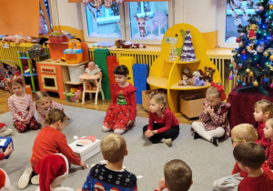 Dzieci siedzą na dywanie, dziewczynka karmi Mikołaja ciasteczkami zgodnie z wylosowaną cyfrą na kostce.