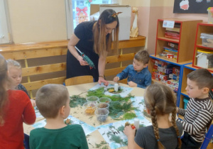 Na zdjęciu widzimy dzieci siedzące przy stoliku które wykonują swoje stroiki, oraz Panią która pomaga przyklejać gałązki choinek do podstawy stroika.
