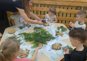 Na zdjęciu widzimy dzieci siedzące przy stoliku które wykonują swoje stroiki, oraz Panią która pomaga przyklejać gałązki choinek do podstawy stroika.