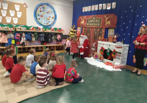 Na zdjęciu widzimy dzieci siedzące na dywanie, w tle widać dekoracje związaną z wizytą Mikołaja oraz Mikołaja wręczającego upominek dziewczynce