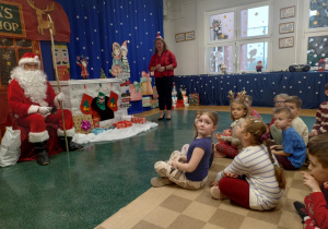 Zdjęcie przedstawia dzieci siedzące na dywanie przed Mikołajem oraz Panią Dyrektor stojącą obok. Dzieci śpiewają piosenkę Mikołajowi