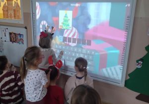 Na zdjęciu widzimy dziewczynkę stojącą przed tablicą interaktywna, która rozwiązuje krzyżówkę związaną z osobą Św. Mikołaja