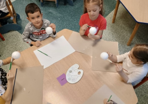 Dzieci siedzą przy stole i wykonują świąteczne prace plastyczne, ozdabiając bombki kolorowymi serwetkami.