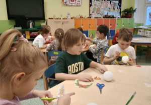 Dzieci siedzą przy stoliku i zdobią bombki styropianowe.