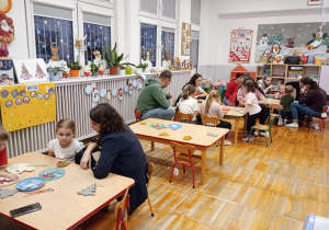 Rodzice wraz z dziećmi siedzą przy trzech ławkach wspólnie wykonując ozdobę choinkowa.