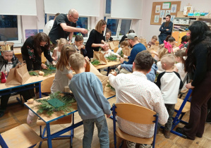 Zdjęcie przedstawia dzieci wraz z ich rodzicami wykonującymi stroik świąteczny.