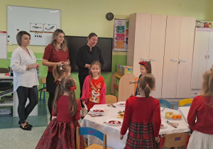 Dzieci stoją przy stołach, a panie składają życzenia.