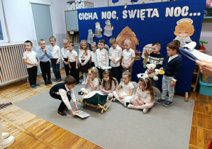 Chłopiec kładzie książkę przed żłóbkiem. Cztery dziewczynki siedzą wokół żłobka. Za nimi stoją przedszkolaki w odświętnych strojach na tle dekoracji.