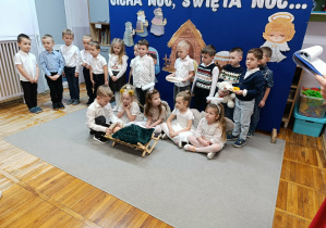 Chłopiec siedzący przy żłóbku zakłada czapkę na głowę lalki, symbolizującej Jezuska. Dziewczynki w strojach aniołków siedzą za żłóbkiem. Pozostałe dzieci stoją na tle dekoracji.