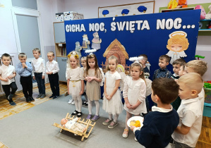 Cztery dziewczynki w strojach aniołków stoją przed żłóbkiem. Za nimi dzieci w odświętnych strojach stoją na tle bożonarodzeniowej dekoracji.