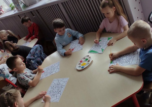 Dzieci siedzące przy stolikach wykonują pracę plastyczną z wykorzystaniem farby.