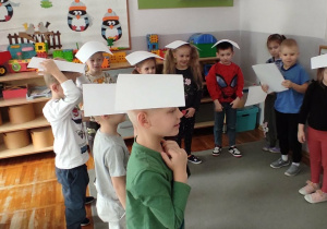 Dzieci stoją na dywanie z białym kartkami na głowach.