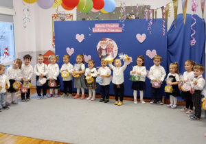 Przedszkolaki stojące w półkolu trzymające w rękach upominki dla babć i dziadków.