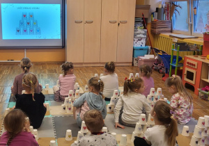 Dzieci siedzą na dywanie, odtwarzają z kubeczków wyświetlony na tablicy kod.