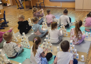 Dzieci siedzą na dywanie, odtwarzają z kubeczków wyświetlony na tablicy kod.