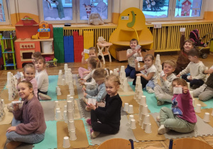 Dzieci siedzą na dywanie, odtwarzają z kubeczków podany na kartoniku kod.