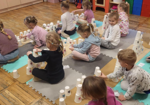 Dzieci siedzą na dywanie, odtwarzają z kubeczków podany na kartoniku kod.