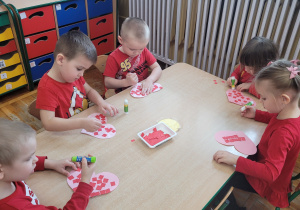 Dzieci z grupy słoneczka siedzą przy stolikach i wyklejają różowe serduszka czerwonymi kwadracikami.