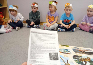 Dzieci siedzą na dywanie i słuchają opowiadania czytanego przez nauczycielkę.