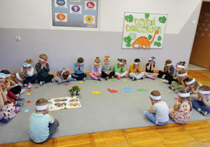 Przedszkolaki siedzą w kole z zamkniętymi oczami w trakcie, gdy nauczycielka rozdaje różnokolorowe papierowe jaja.