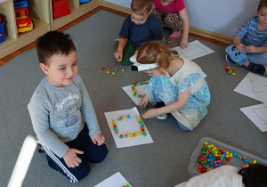 Przedszkolaki siedzą na dywanie i z kolorowych koralików układają kontury figur geometrycznych.