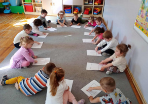 Przedszkolaki siedzą w kole i palcem poprawiają figury narysowane na kartce.