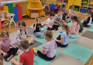 Dzieci siedzą na macie z rękoma na kolanach, uczestniczą w zabawie ,,Kto pierwszy weźmie woreczek”.