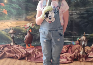 Dziewczynka w stroju ogrodniczki stoi na scenie z mikrofonem w dłoni.