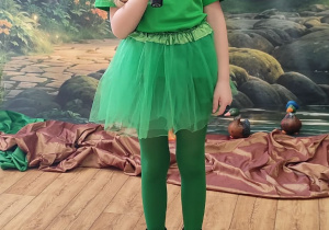 Dziewczynka w przebraniu żaby, trzymająca mikrofon w dłoni śpiewa piosenkę.