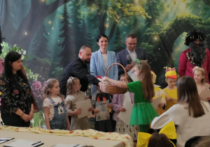 Na scenie stoją dzieci występujące w konkursie wraz z jury, jedna dziewczynka odbiera upominek z koszyka.