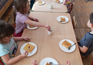 Dzieci siedzą przy stolikach i zjadają upieczone gofry.