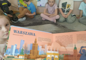 Na zdjęciu widzimy dzieci siedzące na dywanie które słuchają ciekawostek o mieście Warszawie czyli stolicy naszego kraju