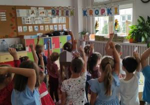 Grupa dzieci stoi w sali przedszkolnej trzymając w wyciągniętych do góry dłoniach kropki. Nad głowami dzieci zawieszony jest napis „Dzień Kropki”.