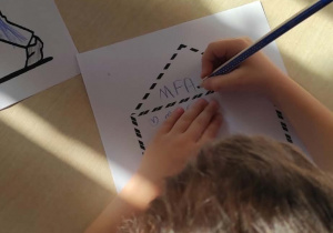 Dziewczynka siedząca przy stoliku próbuje zapisać kredką adres na kopercie.
