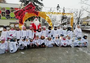 Dzieci przebrane w stroje aniołków pozuja do zdjęcia na podświetlonej ściance z napisem wesołych świąt. Za nimi stoi święty Mikołaj.