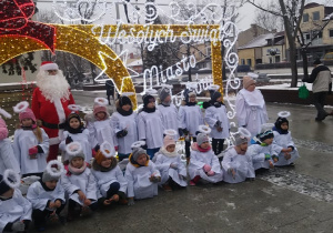 Dzieci przebrane w stroje aniołków pozuja do zdjęcia na podświetlonej ściance z napisem wesołych świąt. Za nimi stoi święty Mikołaj.