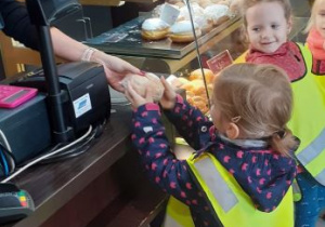 Dziewczynka z grupy Stokrotki stoi przy ladzie w piekarni i kupuje świeżo upieczoną bułkę.