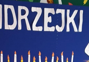 Na granatowym tle napis wykonany bialymi literami - Andrzejki. Pod spodem rząd białych i żółtych świeczek oraz karty do gry.