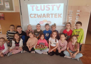 Dzieci z grupy Smerfy pozują do zdjęcia na tle napisu „Tłusty czwartek”. Jedno dziecko trzyma na kolanach tace z pączkami.