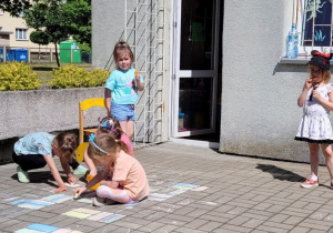 Dzieci z grupy Misie spędziły ten dzień na dworze. Dwie dziewczynki malują kredą na tarasie i dwie dziewczynki jedzą przekąski.